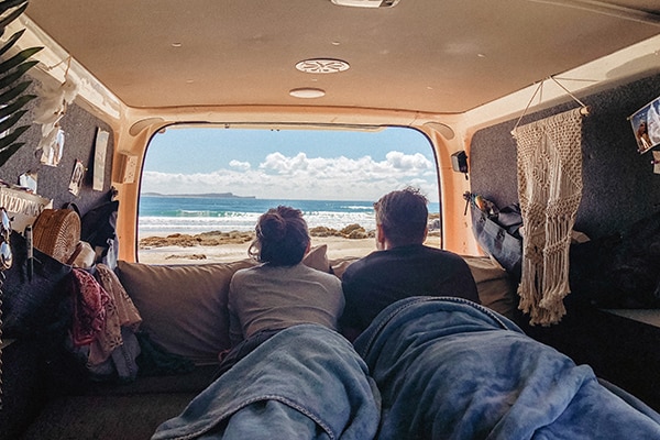 A couple laying in bed looking outside the campervan at the beach